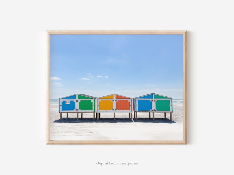 May include: A framed print featuring three colorful beach huts on a sandy shore under a clear blue sky. The huts are painted in vibrant hues of blue, green, orange, and yellow. The image is a coastal photography print.