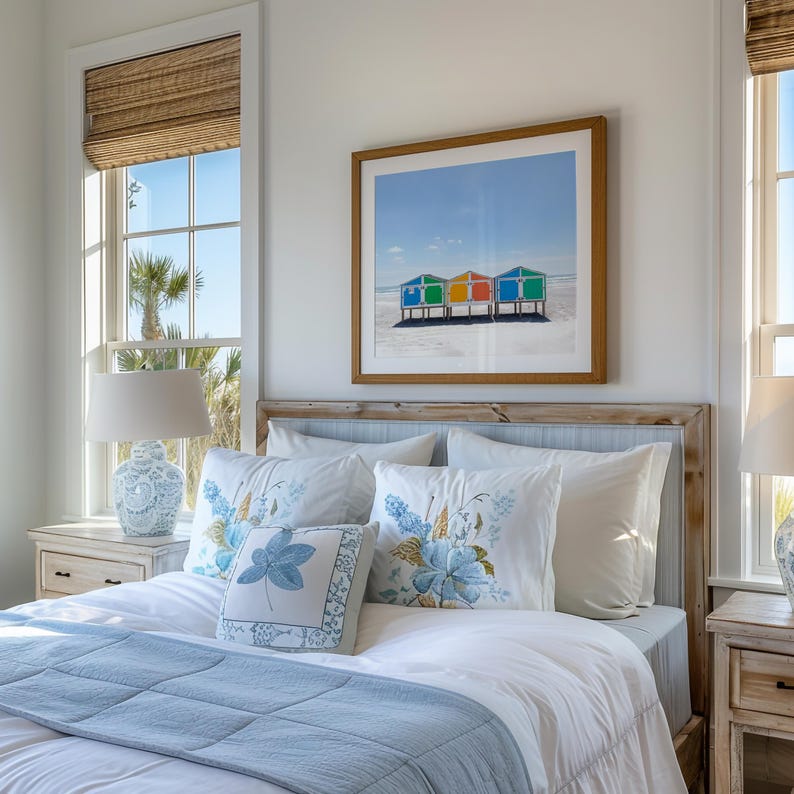 May include: A bedroom scene featuring a bed with white bedding, a light blue quilt, and decorative pillows with floral patterns. A framed print of colorful beach huts hangs above the bed. A lamp sits on a nightstand.