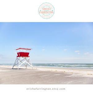 May include: A red and white lifeguard stand on a sandy beach under a clear blue sky. The ocean is visible in the background with small waves. The image has a bright and airy feel.