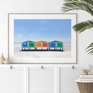 May include: A framed print featuring a beach scene with colorful beach huts. The huts are painted in bright blue, green, yellow, and red, standing on a sandy beach under a clear blue sky. The image evokes a sense of summer and coastal charm.