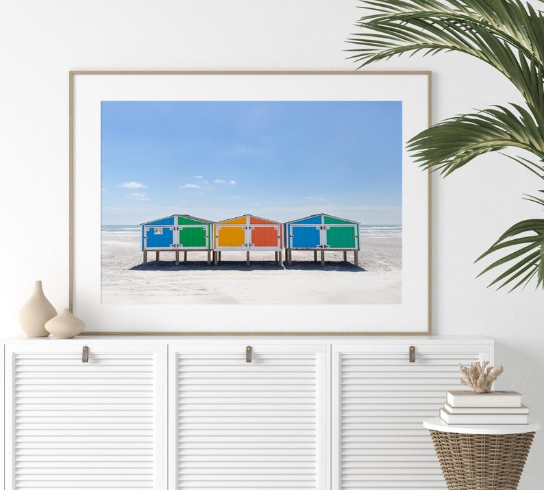 May include: A framed print featuring a beach scene with colorful beach huts. The huts are painted in bright blue, green, yellow, and red, standing on a sandy beach under a clear blue sky. The image evokes a sense of summer and coastal charm.