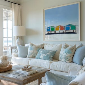 May include: A living room with a white sofa, light blue pillows, and a large framed print of colorful beach huts. A wooden coffee table with books and decorative items completes the coastal theme.