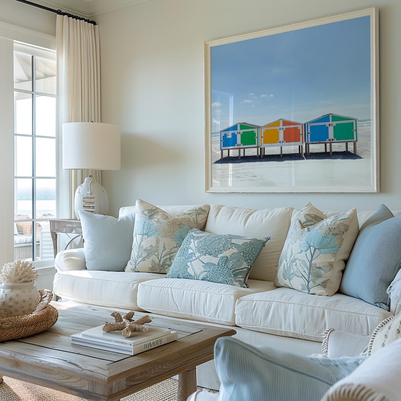 May include: A living room with a white sofa, light blue pillows, and a large framed print of colorful beach huts. A wooden coffee table with books and decorative items completes the coastal theme.