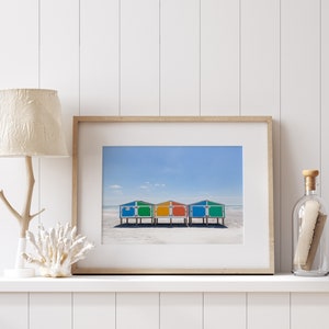 May include: Framed print featuring three colorful beach huts with blue, green, and orange roofs, set against a sandy beach and a clear blue sky. The artwork is displayed on a white shelf with decorative elements.