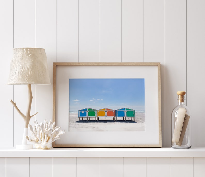May include: Framed print featuring three colorful beach huts with blue, green, and orange roofs, set against a sandy beach and a clear blue sky. The artwork is displayed on a white shelf with decorative elements.