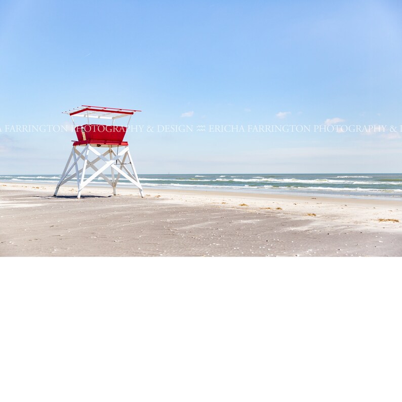 Puede incluir: Un puesto de salvavidas rojo y blanco en una playa de arena con cielo azul y oc&eacute;ano en el fondo.