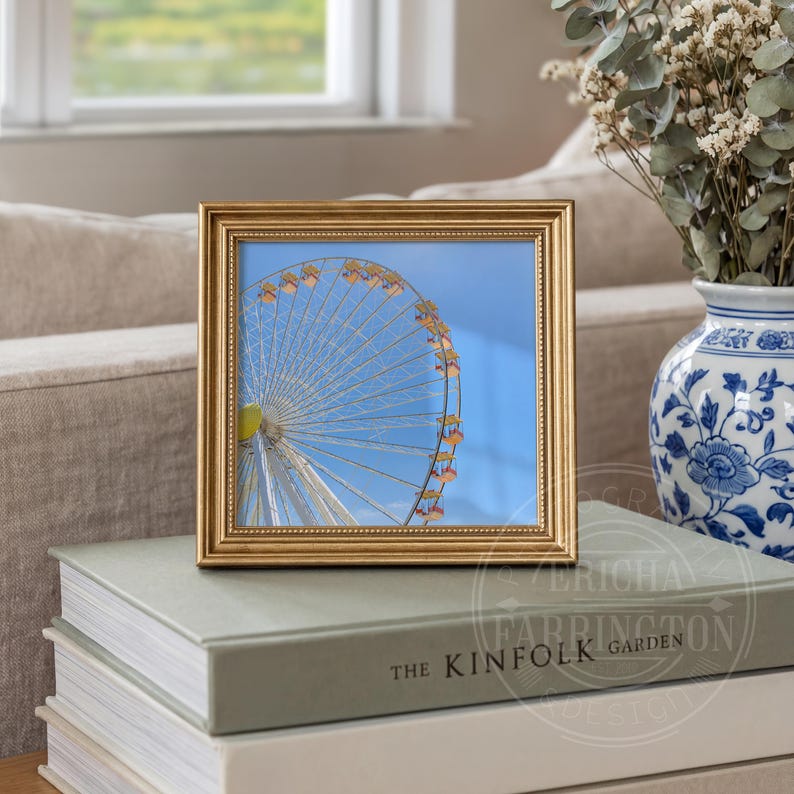 May include: A square, gold-framed picture featuring a Ferris wheel against a bright blue sky. The picture is resting on a stack of books, with a decorative vase and flowers in the background.