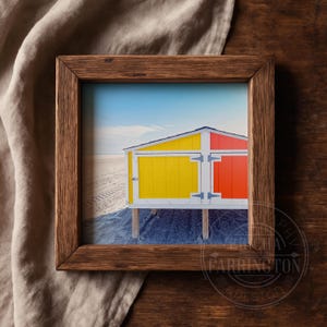 May include: A framed print depicting a beach scene with vibrant beach huts. The huts are painted yellow and red, set on wooden supports against a sandy beach and a blue sky. The frame is made of dark wood.