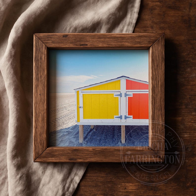 May include: A framed print depicting a beach scene with vibrant beach huts. The huts are painted yellow and red, set on wooden supports against a sandy beach and a blue sky. The frame is made of dark wood.