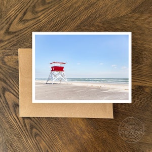 Puede incluir: Un puesto de salvavidas rojo y blanco en una playa de arena con cielo azul y olas del oc&eacute;ano en el fondo.