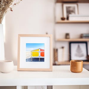 May include: A framed print of a beach hut with yellow and red panels against a blue sky. The artwork is displayed on a white shelf, with a white vase of dried flowers and a small, beige ceramic pot. A wooden cup is also present.