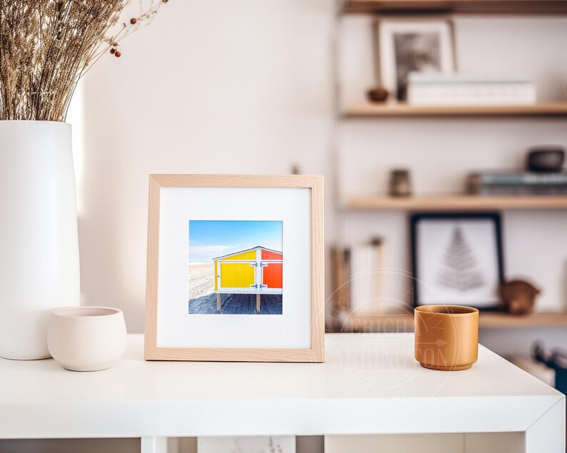 May include: A framed print of a beach hut with yellow and red panels against a blue sky. The artwork is displayed on a white shelf, with a white vase of dried flowers and a small, beige ceramic pot. A wooden cup is also present.