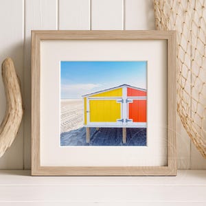 May include: Framed print featuring a vibrant beach hut. The hut is split into yellow and orange panels with a white door and black hinges. The background shows a sandy beach and a clear blue sky.