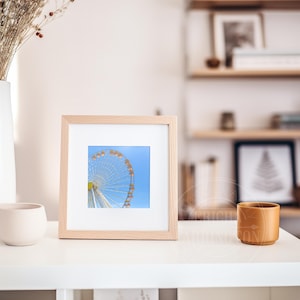 May include: A framed print featuring a partial view of a Ferris wheel against a bright blue sky. The print is in a light wood frame, displayed on a white surface. A white vase with dried flowers and a small beige pot are also on the surface.