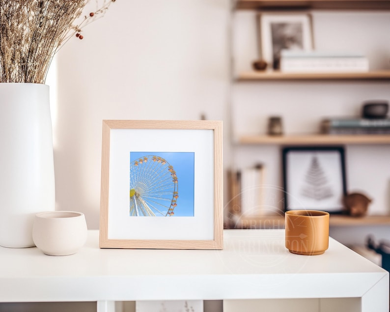 May include: A framed print featuring a partial view of a Ferris wheel against a bright blue sky. The print is in a light wood frame, displayed on a white surface. A white vase with dried flowers and a small beige pot are also on the surface.