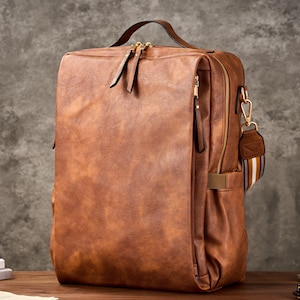 Może przedstawiać: Brązowy skórzany plecak z górnym uchwytem i wieloma przegrodami zapinanymi na zamek. Torba ma prostokątny kształt i pasek na ramię w paski. Laptop i inne przedmioty znajdują się na drewnianej powierzchni.