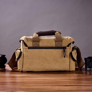 May include: A tan canvas camera bag with brown leather accents and a zippered closure. The bag has a shoulder strap and is sitting on a wooden surface with two camera lenses.