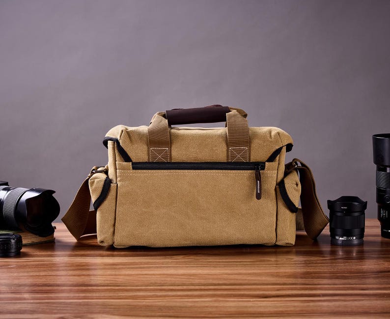 May include: A tan canvas camera bag with brown leather accents and a zippered closure. The bag has a shoulder strap and is sitting on a wooden surface with two camera lenses.