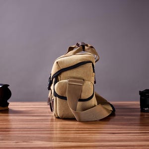 May include: A tan canvas camera bag with a brown leather strap and black accents. The bag is sitting on a wooden table with two camera lenses in the background.