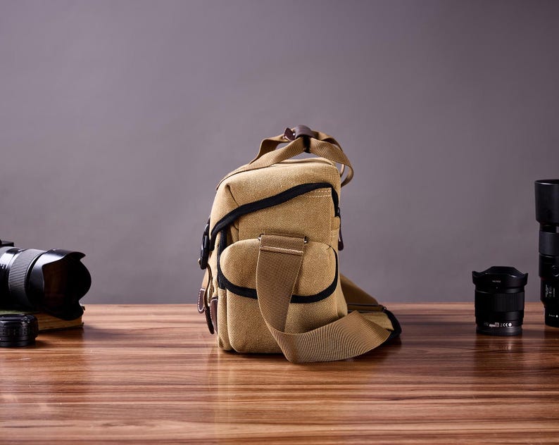 May include: A tan canvas camera bag with a brown leather strap and black accents. The bag is sitting on a wooden table with two camera lenses in the background.