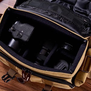 May include: A tan canvas camera bag with a zippered top and adjustable shoulder strap. The bag is open and contains a black camera, a lens, and other camera accessories.