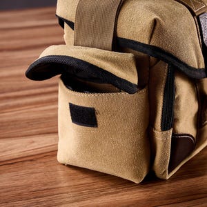 May include: A tan canvas camera bag with brown leather accents and black straps. The bag has a flap that folds over the top and a zippered pocket on the front.