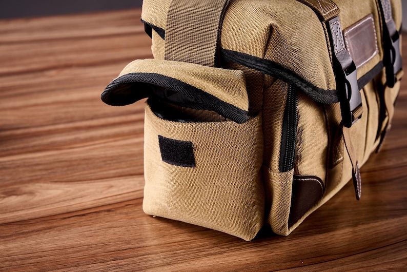 May include: A tan canvas camera bag with brown leather accents and black straps. The bag has a flap that folds over the top and a zippered pocket on the front.