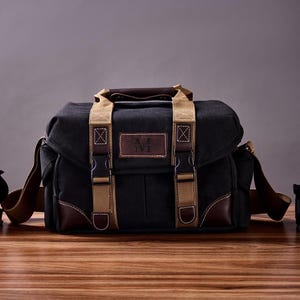 May include: A black canvas camera bag with brown leather accents and tan straps. The bag has a metal buckle and a logo that says "N.K. NYC".