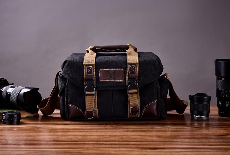 May include: A black canvas camera bag with brown leather accents and tan straps. The bag has a metal buckle and a logo that says "N.K. NYC".