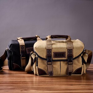 May include: Two camera bags, one black and one tan, with adjustable straps and a leather patch on the front. The tan bag has the initials "RHK" on the patch.
