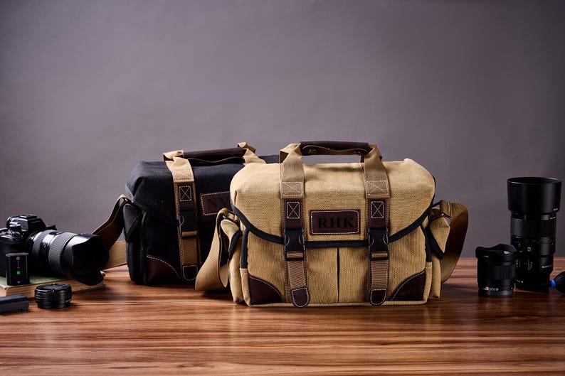 May include: Two camera bags, one black and one tan, with adjustable straps and a leather patch on the front. The tan bag has the initials "RHK" on the patch.