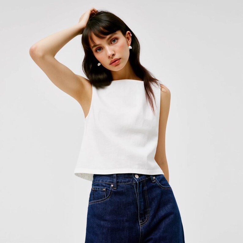 May include: A white sleeveless top with a square neckline and a cropped fit. The top is made of a lightweight fabric and has a relaxed fit. The model is wearing the top with dark blue jeans.