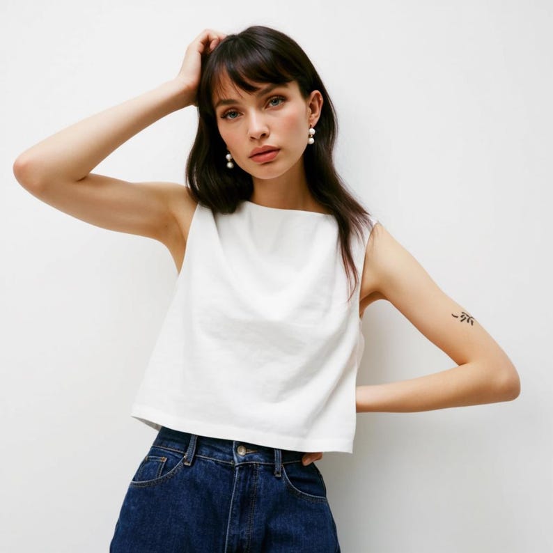 May include: A white sleeveless cropped top with a square neckline. The top is made of a soft, lightweight fabric and has a relaxed fit. The model is wearing the top with blue jeans.