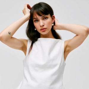 May include: A woman with dark hair wearing a white sleeveless top and pearl earrings.