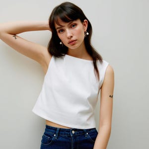 May include: A woman wearing a white sleeveless crop top and blue jeans. She has dark hair and is wearing pearl earrings.