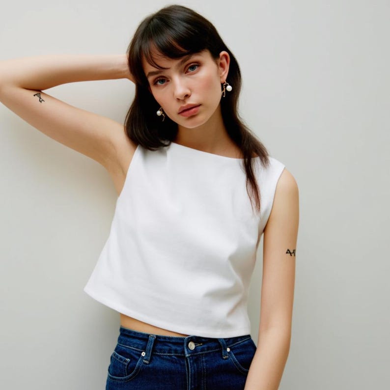 May include: A woman wearing a white sleeveless crop top and blue jeans. She has dark hair and is wearing pearl earrings.