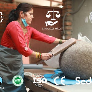 May include: A woman in a red shirt and grey skirt is working on a large, round, grey felt ball. She is using a wooden tool to shape the ball. The image includes several certifications, including Fairtrade, GMP Quality, ISO, CE, and Sedex. The text "100% Original", "Ethically Made", and "Handmade with Love" is also visible.