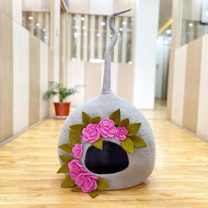 以下が含まれることがあります： 長い尻尾が付いた花瓶の形をした灰色のフェルト製の猫用ベッド。ベッドの開口部にはピンクの花の模様があります。