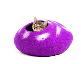 May include: A purple felt cat bed with white flecks. A tabby cat is peeking out of the opening of the bed.