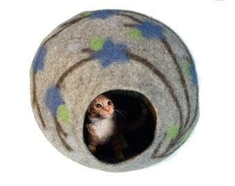 Könnte beinhalten: Ein graues Filzkatzenbett mit einem blauen und grünen Blumendesign. Eine kleine orange und weiße Katze schaut aus dem Eingang.