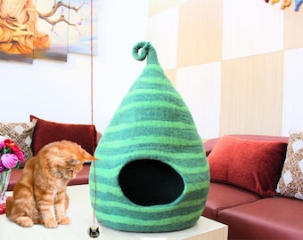 Puede incluir: Una cama para gatos de color verde y blanco a rayas con forma de lágrima y una parte superior enroscada. Un gato está sentado en el suelo junto a la cama, mirando un juguete en una cuerda.