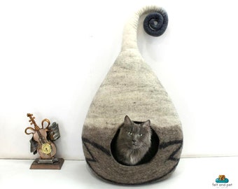 Puede incluir: Un gato gris se sienta dentro de una cama para gatos de fieltro gris y blanco con forma de lágrima y una cola enroscada. La cama tiene una abertura redonda para que el gato pueda entrar.