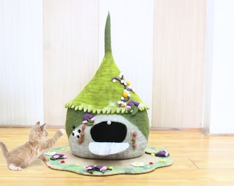 Puede incluir: Una casa para gatos de fieltro verde con forma de techo puntiagudo con una pequeña entrada. La casa está decorada con flores y enredaderas. Un pequeño gatito marrón claro está de pie frente a la casa.