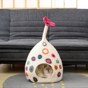 May include: A white felt cat bed with a pink flower on top. The bed is decorated with colorful circles and has a round opening for the cat to enter. A cat is sitting inside the bed.
