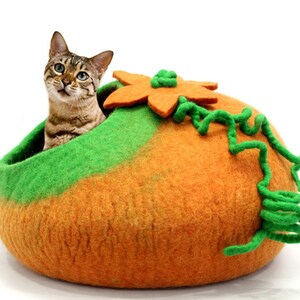 以下が含まれることがあります： 緑とオレンジのフェルト製の猫用ベッドで、かぼちゃの形をしています。緑のフェルトの茎と花が上についています。猫がベッドの中に座っています。