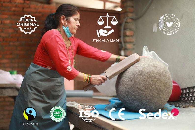 May include: A woman working on a large, round, gray felt ball with a wooden tool. She is wearing a red shirt and a gray apron. The image includes text that says "100% Original", "Ethically Made", "Handmade with Love", "Fairtrade", "GMP Quality", "ISO", "CE", and "Sedex".