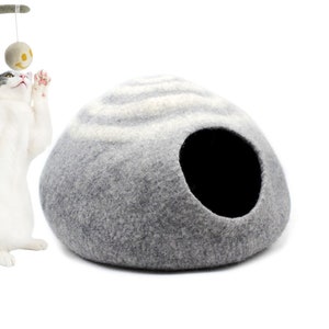 May include: A grey and white felt cat bed shaped like a dome with a round opening. A white cat with grey markings is playing with a toy on a stick.