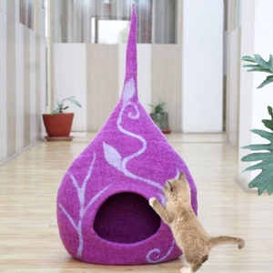 May include: A purple felt cat bed shaped like a teardrop with a white floral design. A cat is standing near the entrance of the bed.