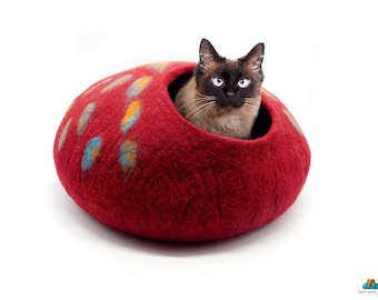 May include: A red felt cat bed with blue, yellow, and orange spots. A brown and black cat is peeking out of the bed.
