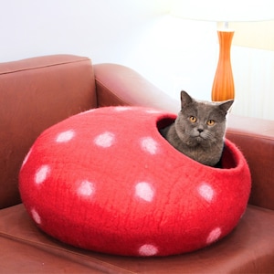 Puede incluir: Un gato gris descansando dentro de una cama para gatos redonda de fieltro rojo con lunares blancos. La cama para gatos está en un sofá de cuero marrón.